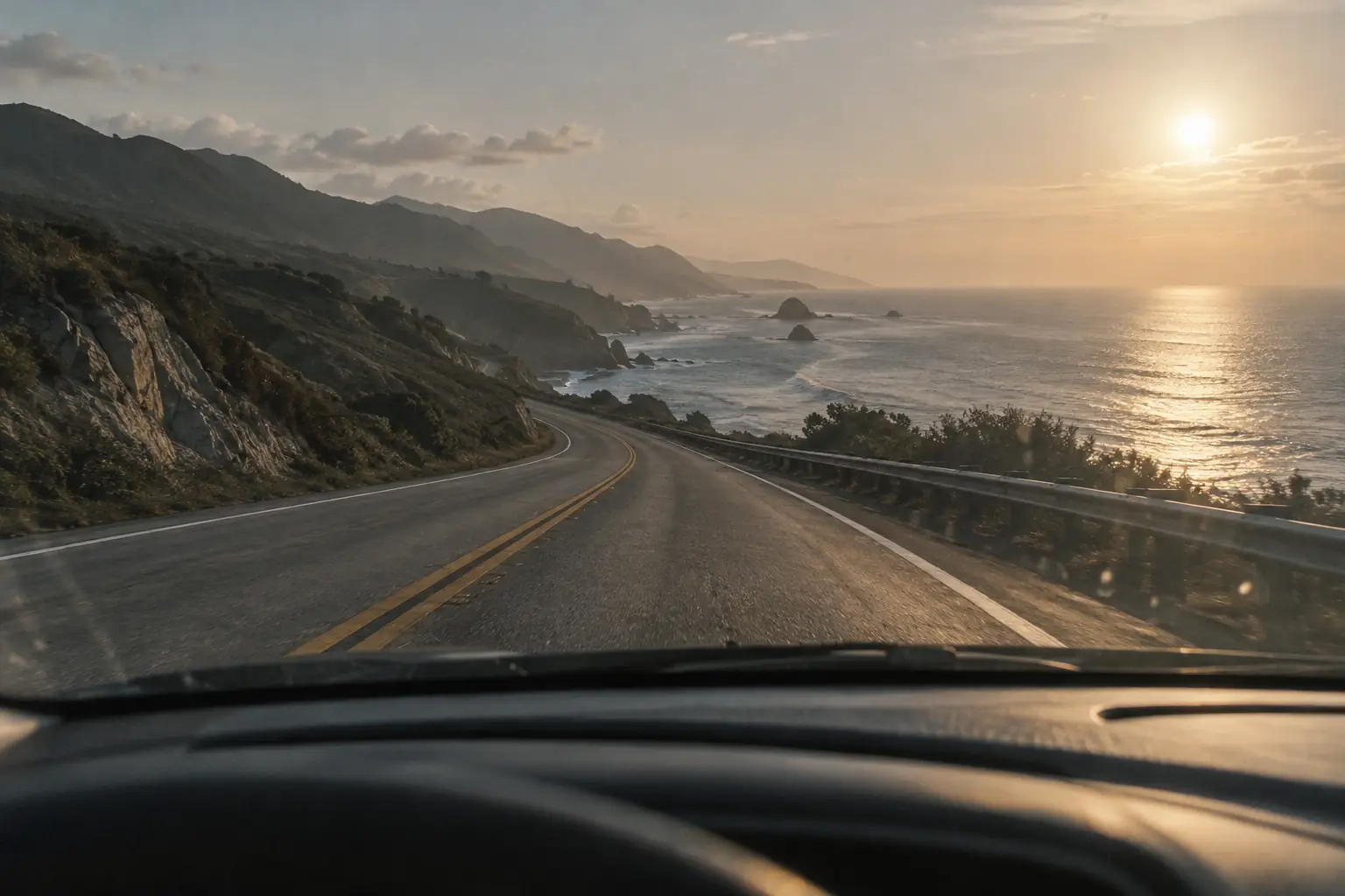 An empty coastal highway seen through a car windshield in late afternoon light.